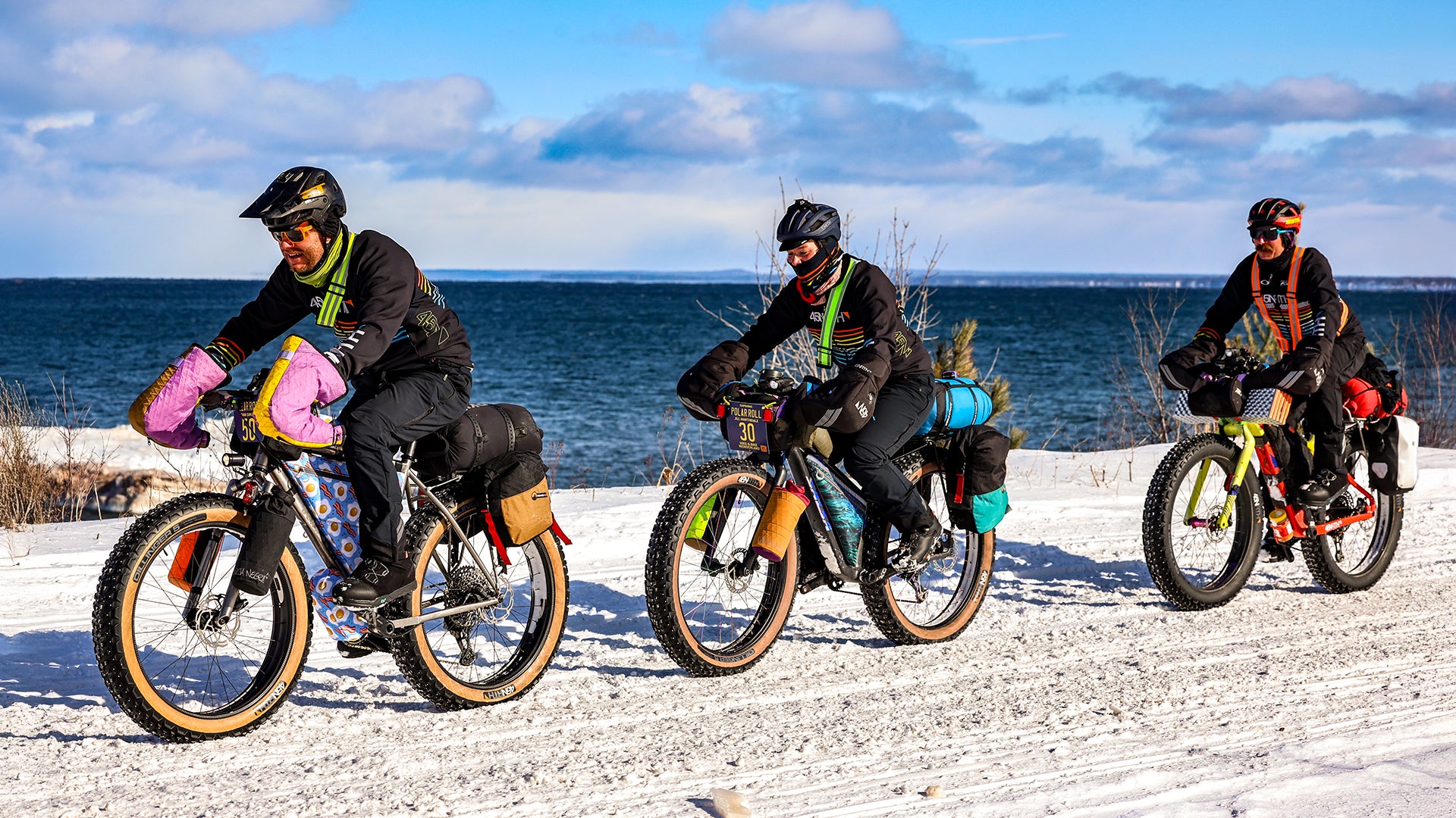 Three fat bikers riding next to snowy lake shore with fully loaded bikes