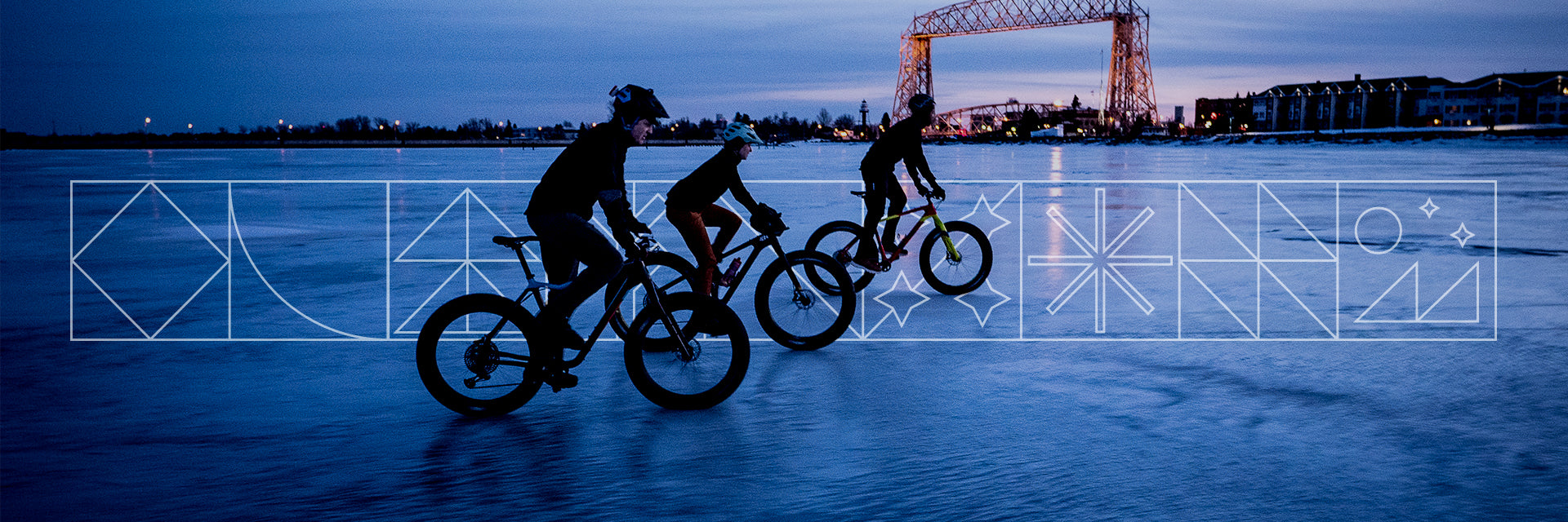Three fat bikers riding on frozen lake at dusk lift bridge in background