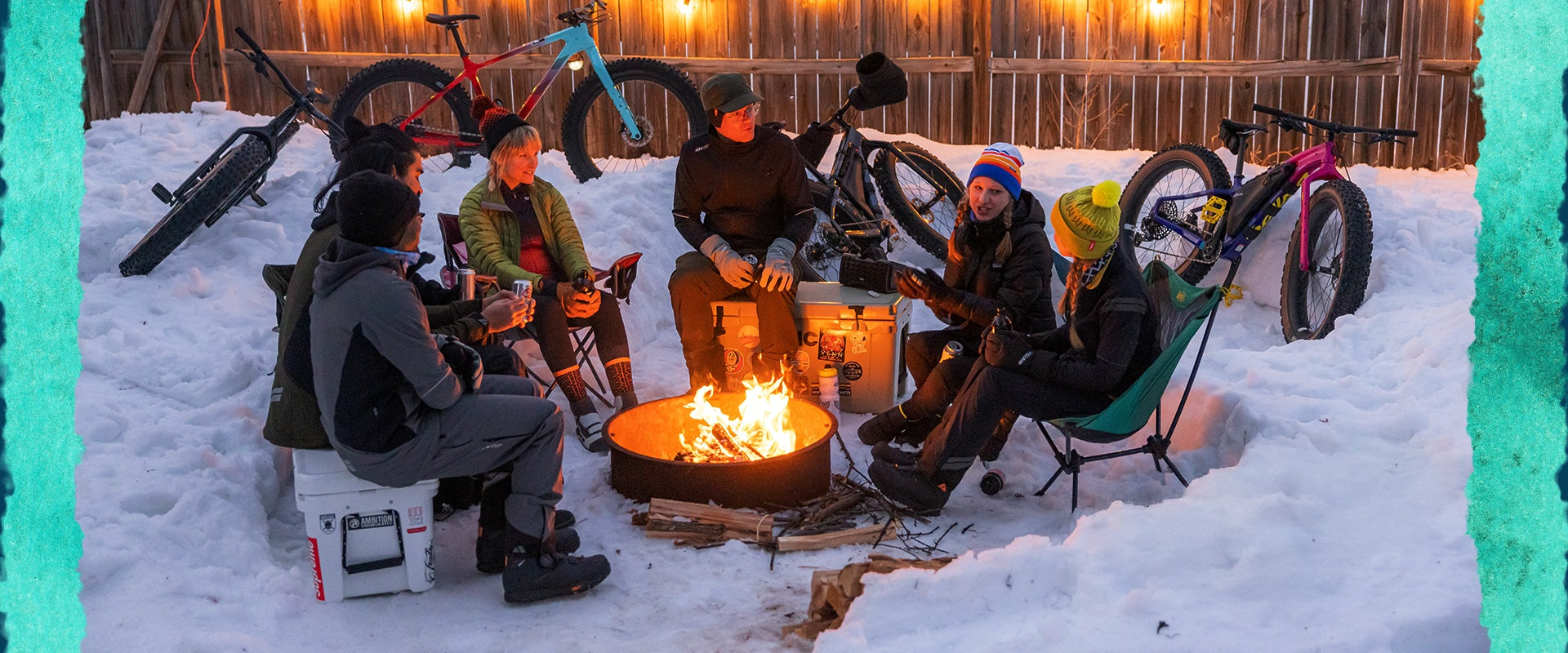 Group of people sitting around a campfire in the snow with bicycles nearby.